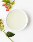 A white bowl filled with pale Mountain Rose Herbs Rosehip Seed Oil sits on a white surface beside sprigs of greenery with small, round red berries.
