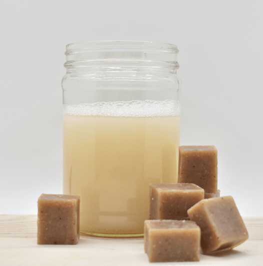 A glass jar of Tangie Liquid Laundry Concentrate sits on a light surface, with small brown laundry soap cubes beside it. This plastic-free detergent is gentle for sensitive skin. The background is plain white.