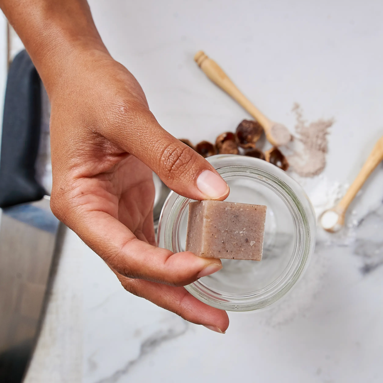 A hand holds a small glass jar of Tangie Liquid Laundry Concentrate. On the white surface below are a wooden spoon, honey dipper, powder, and round brown objects—ideal for plastic-free laundry routines.