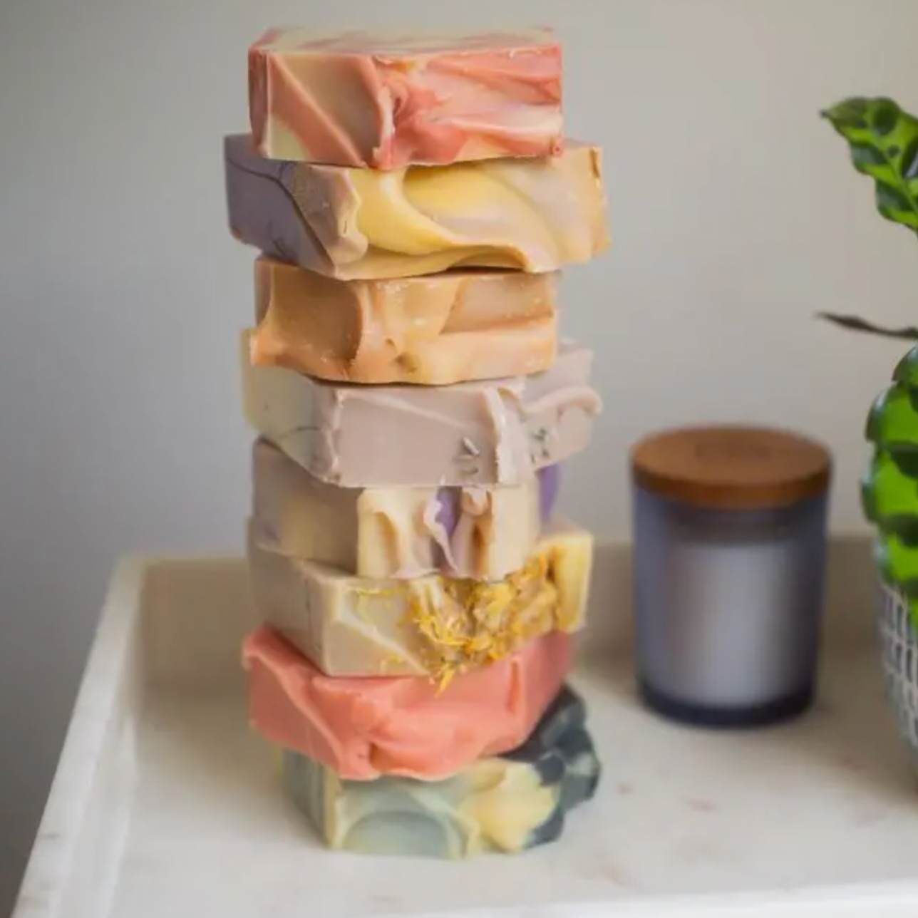 A stack of seven colorful Perennial Soaps Bar Soaps is arranged on a white surface, with a potted plant and a candle with a wooden lid in the background.