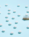 Huppy Kids Toothtabs—white round tablets—are scattered on a light blue surface next to a wooden toothbrush on the right.