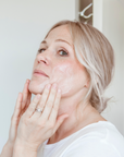 A woman with light blonde hair, wearing a white shirt in a bright indoor setting, applies Larsons General Brilliant Bamboo Exfoliating Face Wash to her face with both hands as she looks slightly upward.