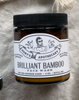 A brown jar labeled Larsons General Brilliant Bamboo Exfoliating Face Wash with a black lid sits on beige fabric. The label highlights bamboo powder, product details, and features a woman in sunglasses illustration.