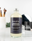 A clear Zerra & Co. Makeup Remover bottle, ideal for sensitive skin, sits on a white surface with makeup brushes, waterproof mascara, a mirror, and foundation products blurred in the background.