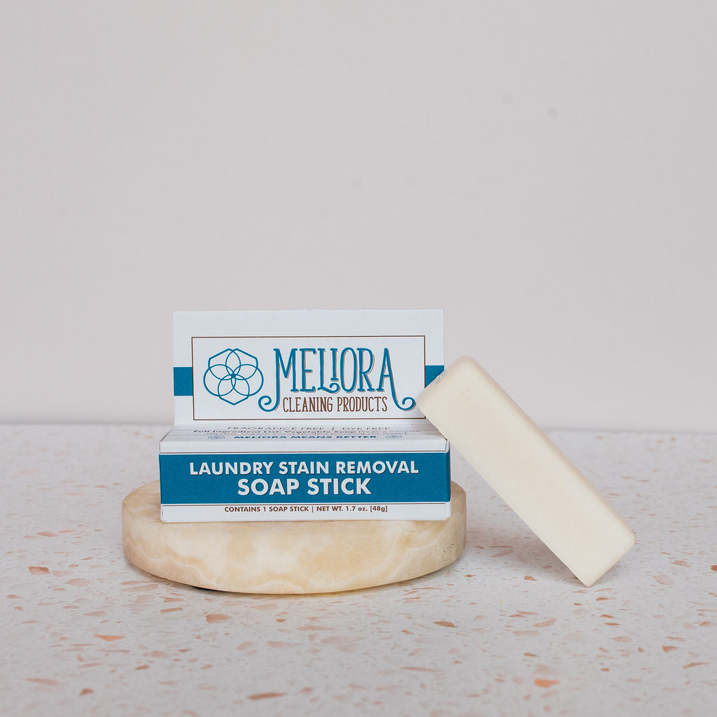 A Meliora Laundry Stain Stick stands upright on a round stone tray beside another stick. The white and blue packaging shows product details, while the neutral speckled background creates a clean aesthetic.