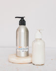 Two pump bottles of All Good Brand are side by side on a cream-colored circular surface: one labeled Sport Sunscreen Lotion SPF 30 and one clear with white lotion, set against a light background.