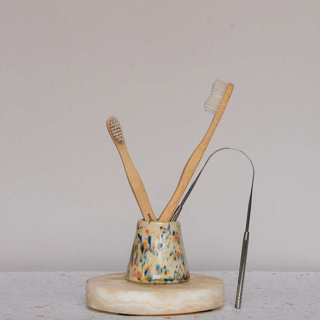 Two Brush With Bamboo Toothbrushes with soft bristles and a metal tongue scraper sit in a speckled ceramic holder on a round light stone, offering an elegant, eco-friendly routine with Bamboo Toothbrush from Brush With Bamboo.