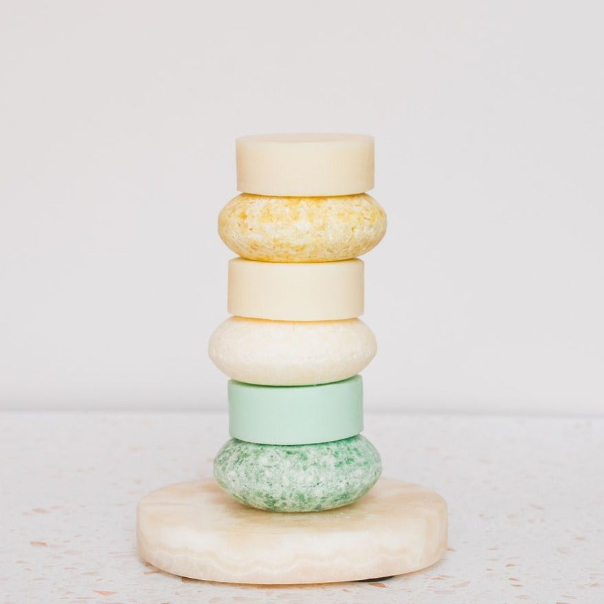 A vertical stack of five Notice Shampoo & Conditioner Bars by Notice Hair Co. in pastel yellow, white, green, and cream sits on a round stone base. These bars are perfect for color-treated hair and displayed against a light background.