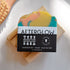 A vibrant Perennial Soaps Bar Soap with yellow, teal, and pink swirls rests on a wooden dish. The AFTERGLOW label notes its Wisconsin-made, vegan, palm oil free, and handcrafted.