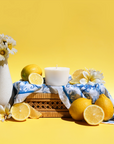 The Arbor Made Candle Refill in the scent Amalfi Lemon. The candle refill is unpackaged in the center surrounded by Lemons and white flowers on a yellow background. 