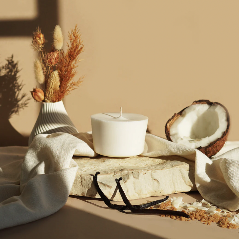 The Arbor Made Candle Refill in the scent Vanilla Colada. The candle refill is unpackaged in the center surrounded by Coconut, vanilla beans, and dried flowers on a tan background. 