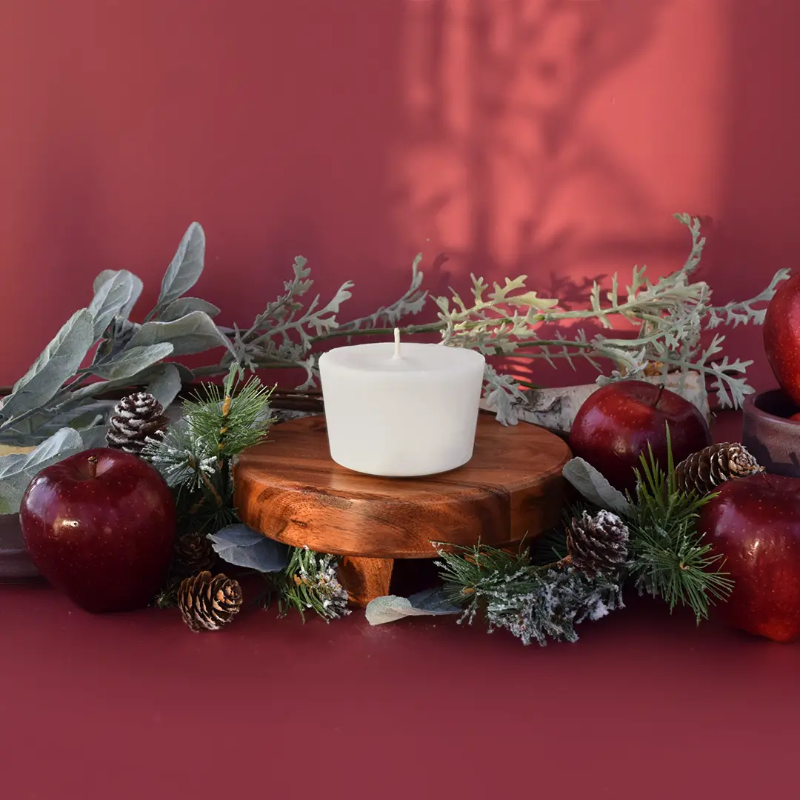 The Arbor Made Candle Refill in the scent Midnight Orchard. The candle refill is unpackaged in the center surrounded by pine decor and apples on a red background. 