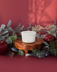 The Arbor Made Candle Refill in the scent Midnight Orchard. The candle refill is unpackaged in the center surrounded by pine decor and apples on a red background. 