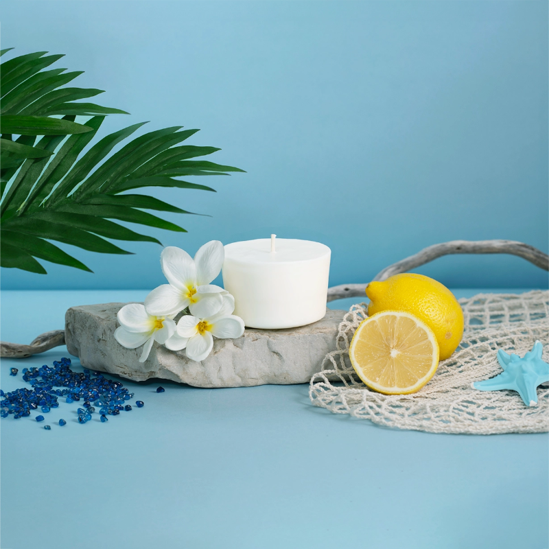 The Arbor Made Candle Refill in the scent Mellow Breeze. The candle refill is unpackaged in the center surrounded by  Lemons, white flowers, and beach decor on a blue background. 