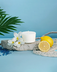 The Arbor Made Candle Refill in the scent Mellow Breeze. The candle refill is unpackaged in the center surrounded by  Lemons, white flowers, and beach decor on a blue background. 