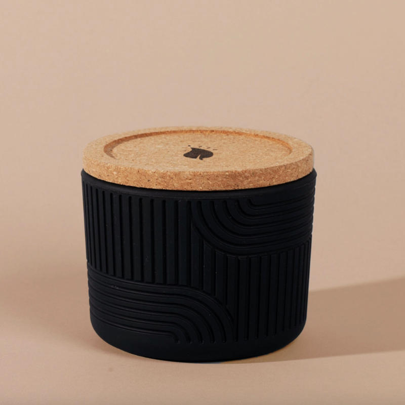 The Arbor Made Refillable Candle Jar in the silicone jar style and in the color black. The jar features a cork lid with a small leaf design on it and a silicone body with a linear geometric pattern on it. The Jar is siting on a beige background. 