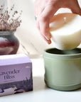 A hand places a white wax refill into an Arbor Made Refillable Candle Jar with a green silicone bottom. Beside it is a purple Arbor Made Lavender Bliss Candle Refill box, with a vase of dried lavender in the background.