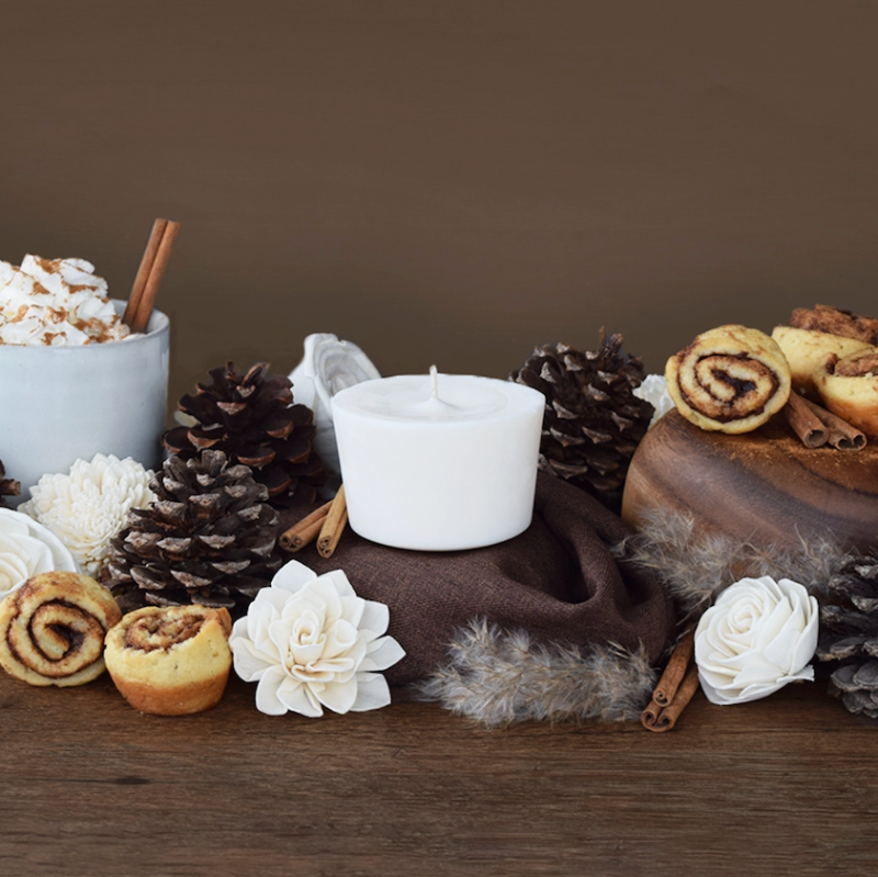The Arbor Made Candle Refill in the scent Cinnamon Swoon. The candle refill is unpackaged in the center surrounded by pine cones, cinnamon sticks, and cinnamon rolls on a brown background. 