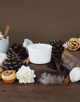 The Arbor Made Candle Refill in the scent Cinnamon Swoon. The candle refill is unpackaged in the center surrounded by pine cones, cinnamon sticks, and cinnamon rolls on a brown background. 