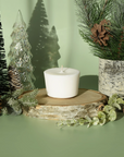 The Arbor Made Candle Refill in the scent Glacial Pine. The candle refill is unpackaged in the center surrounded by pine decor on a green background. 