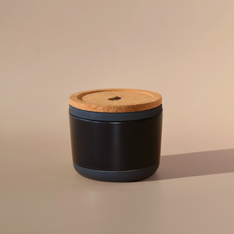 The Arbor Made Refillable Candle Jar by Arbor Made is a small, round black jar with a cork lid and silicone bottom. It sits on a beige surface, casting a soft shadow, while the neutral background highlights its elegant design.
