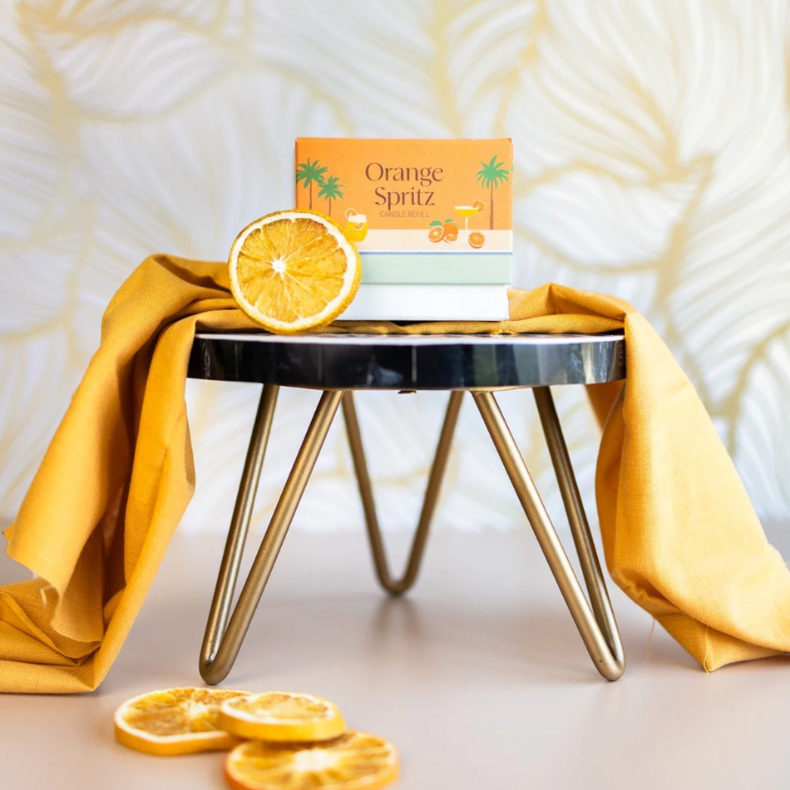 The Arbor Made Candle Refill in the scent Orange Spritz. The candle refill is packaged in the center on a stool surrounded by dried orange slices and orange fabric on a tan background. 