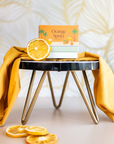 The Arbor Made Candle Refill in the scent Orange Spritz. The candle refill is packaged in the center on a stool surrounded by dried orange slices and orange fabric on a tan background. 