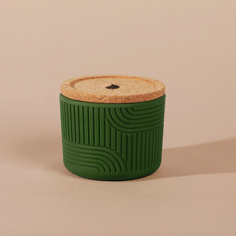 The Arbor Made Refillable Candle Jar in the silicone jar style and in the color green. The jar features a cork lid with a small leaf design on it and a silicone body with a linear geometric pattern on it. The Jar is siting on a beige background. 