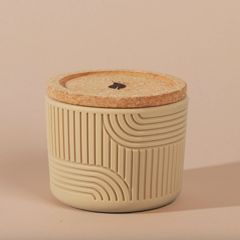 The Arbor Made Refillable Candle Jar in the silicone jar style and in the color sand. The jar features a cork lid with a small leaf design on it and a silicone body with a linear geometric pattern on it. The Jar is siting on a beige background. 