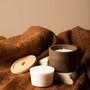 A brown reusable silicone candle jar sits on a stone with a candle burning inside. Next to the jar is a candle refill. 