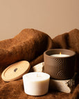 A brown reusable silicone candle jar sits on a stone with a candle burning inside. Next to the jar is a candle refill. 