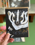 A hand holding a card featuring a high quality print of an oil painting of a badger. 