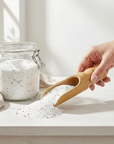A hand holding the Bamboo Scoop and scooping white speckled powder out of a glass jar on a white counter top. 