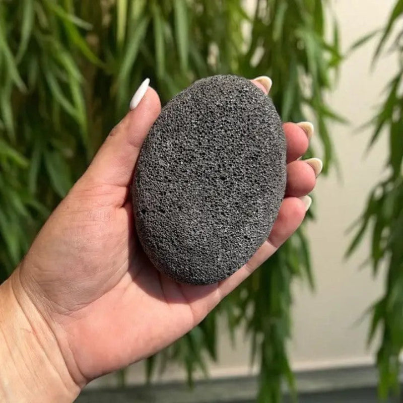 A hand holding a dark grey pumice stone made of natural pumice on a green background.