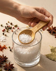 Hand holding a bamboo measuring spoon over a glass of salt with spices and herbs on a beige surface