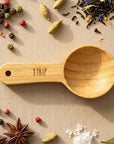 Bamboo measuring spoon labeled '1 TBSP' surrounded by various spices on a beige surface