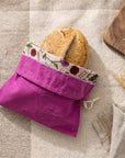 A purple bee's wax lined bread bag with a loaf of bread in it sitting on a linen beige background. A wooden cutting board with bread crumbs on it peeks into frame from the corner.