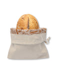 A beige bread bag with a drawstring closure, shown holding a round loaf of bread on a plain white background. The opening is folded down showing its bee's wax lining printed with baked goods illustrations. 
