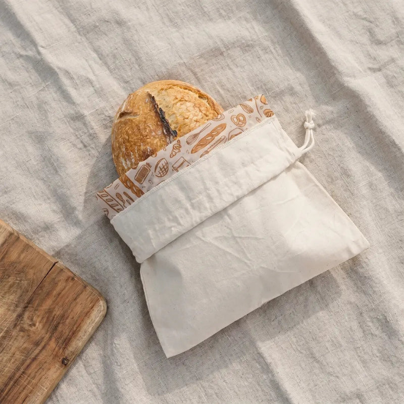 A Loaf of bread in a beige drawstring  bread bag on a linen background.  The reusable bread bag is folded down to show the bee's wax lining printed with illustrations of baked goods. 