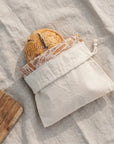 A Loaf of bread in a beige drawstring  bread bag on a linen background.  The reusable bread bag is folded down to show the bee's wax lining printed with illustrations of baked goods. 