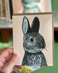 A Hand holding a light pink card featuring a high quality print of an oil painting of a bunny. 