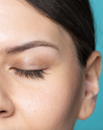 Close-up of a person's face with closed eyes against a teal background. They are wearing the cheekbone mini eyeshadow in the shade brown topaz.