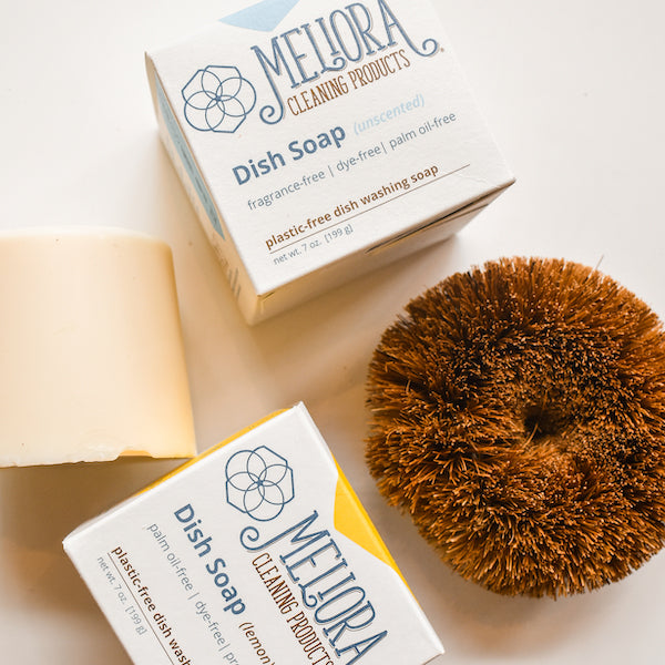 A boxed Meliora Dishwashing Puck and an unwrapped vegan dish soap bar sit beside a brown fiber dish brush. The packaging notes the Meliora Dishwashing Puck is fragrance-free, dye-free, plastic-free, and MADE SAFE Certified.