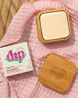 Dip shampoo bar travel case opened up with a shampoo bar inside on a pink fabric background.