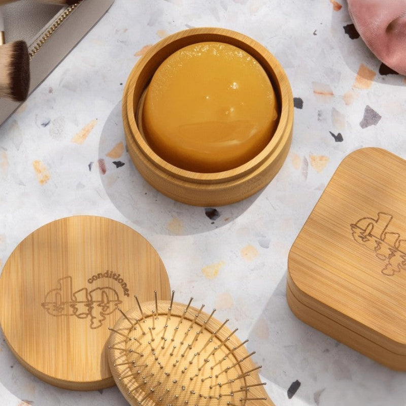 The Dip Large conditioner bar travel case opened up with a conditioner bar inside and on a counter by a brush and the shampoo bar travel case.