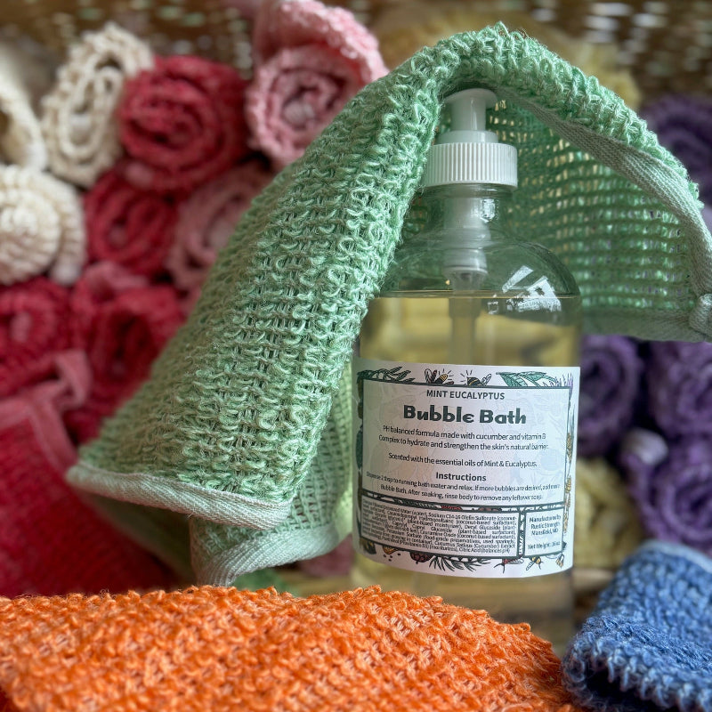 A bottle of Green Life Bubble bath with a green hemp exfoliating washcloth draped over it and a basket full of hemp exfoliating washcloths in the background. 