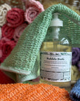 A bottle of Green Life Bubble bath with a green hemp exfoliating washcloth draped over it and a basket full of hemp exfoliating washcloths in the background. 