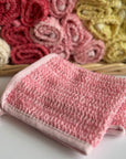 A pink hand-dyed hemp exfoliating washcloth with a basket full of rolled hemp exfoliating washcloths in the background. 
