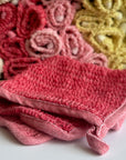 A red hand-dyed hemp exfoliating washcloth with a basket full of rolled hemp exfoliating washcloths in the background. 