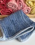 A blue hand-dyed hemp exfoliating washcloth with a basket full of rolled hemp exfoliating washcloths in the background. 
